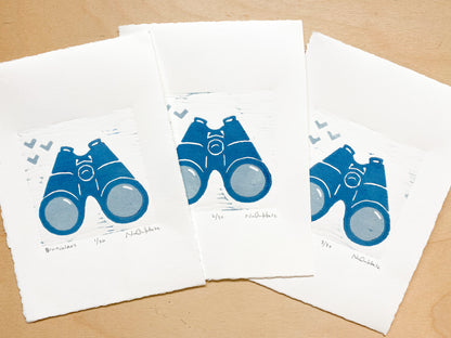 An edition of woodcut prints by Nan Onkka Prints. The prints depict a pair of binoculars printed in two colors - light gray and dark blue. The image is minimalist in style with a hand-carved aesthetic. Made in Grand Marais, Minnesota.