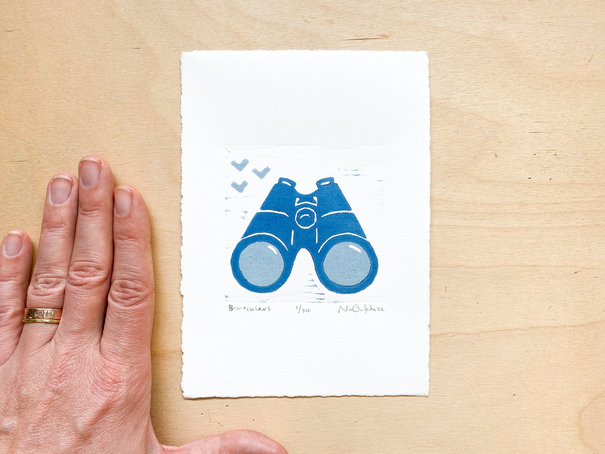 A woodcut print by Nan Onkka Prints. The print depicts a pair of binoculars printed in two colors - light gray and dark blue. The image is minimalist in style with a hand-carved aesthetic. Made in Grand Marais, Minnesota.