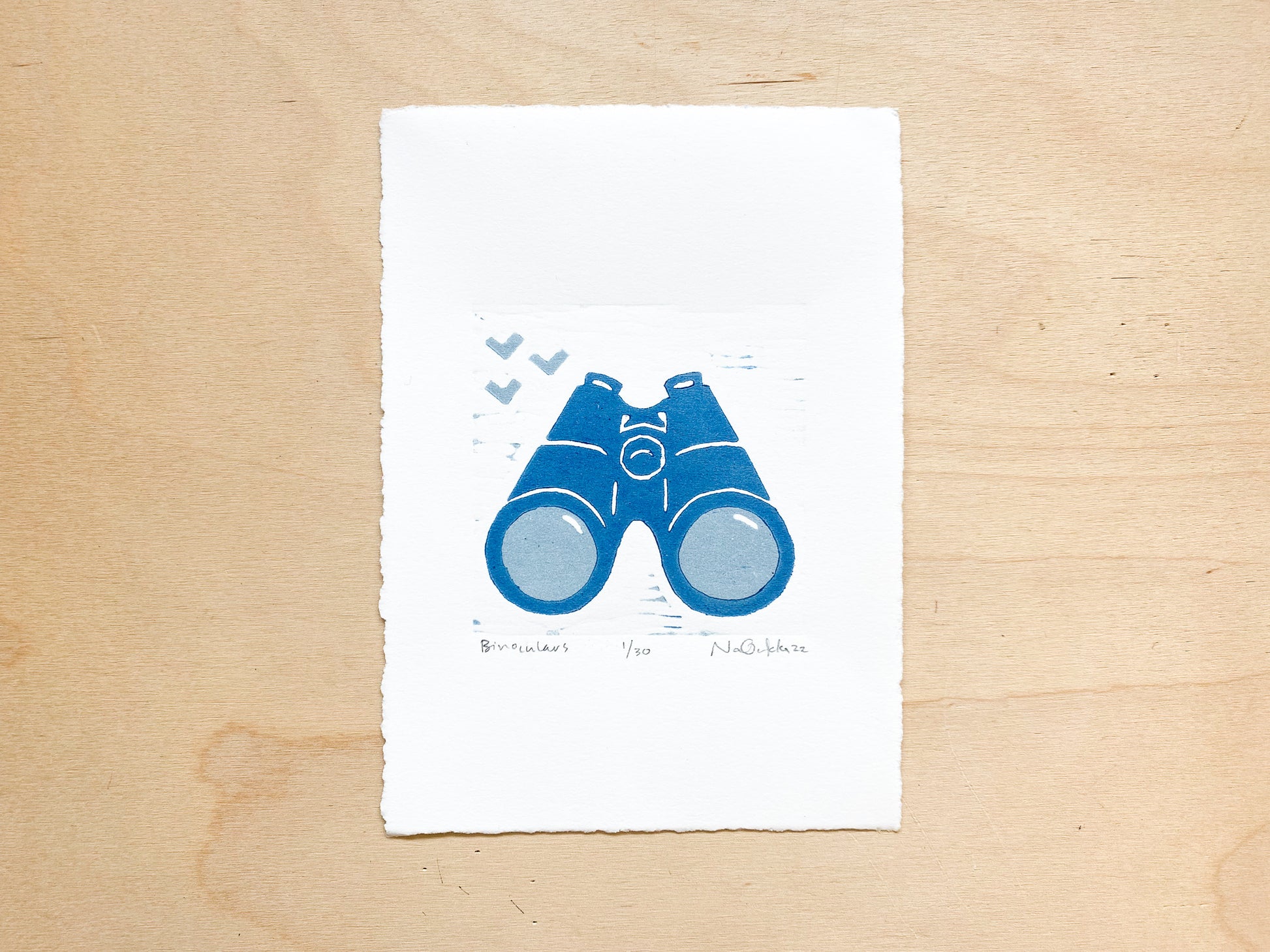 A woodcut print by Nan Onkka Prints. The print depicts a pair of binoculars printed in two colors - light gray and dark blue. The image is minimalist in style with a hand-carved aesthetic. Made in Grand Marais, Minnesota.