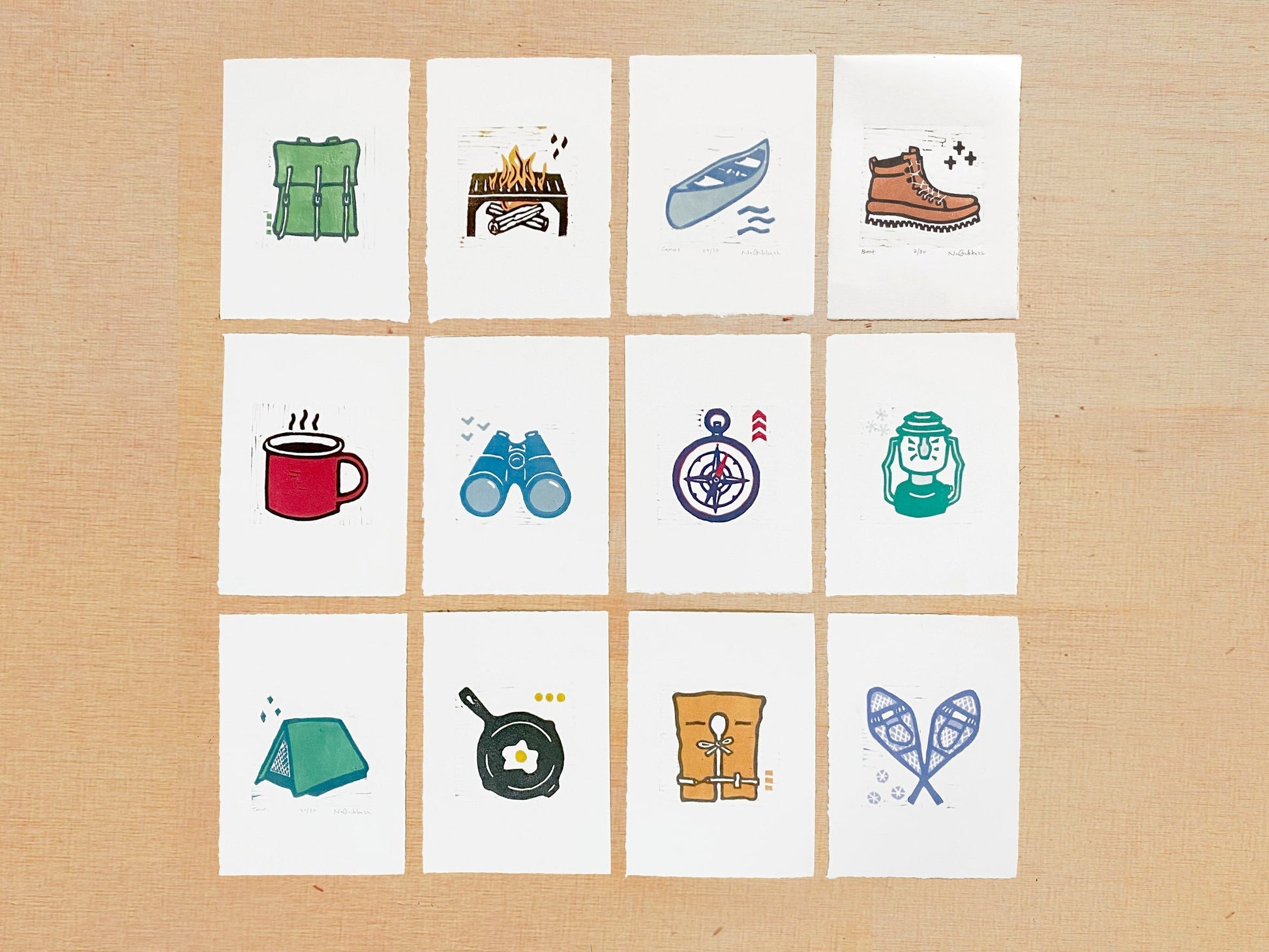 A series of twelve woodcut prints made by Nan Onkka Prints, a printmaking artist in Grand Marais, Minnesota. Each print depicts a different adventure-themed image, including a canoe pack, campfire, canoe, hiking boot, coffee mug, binoculars, compass, lantern, tent, skillet with an egg, life jacket, and snowshoes.