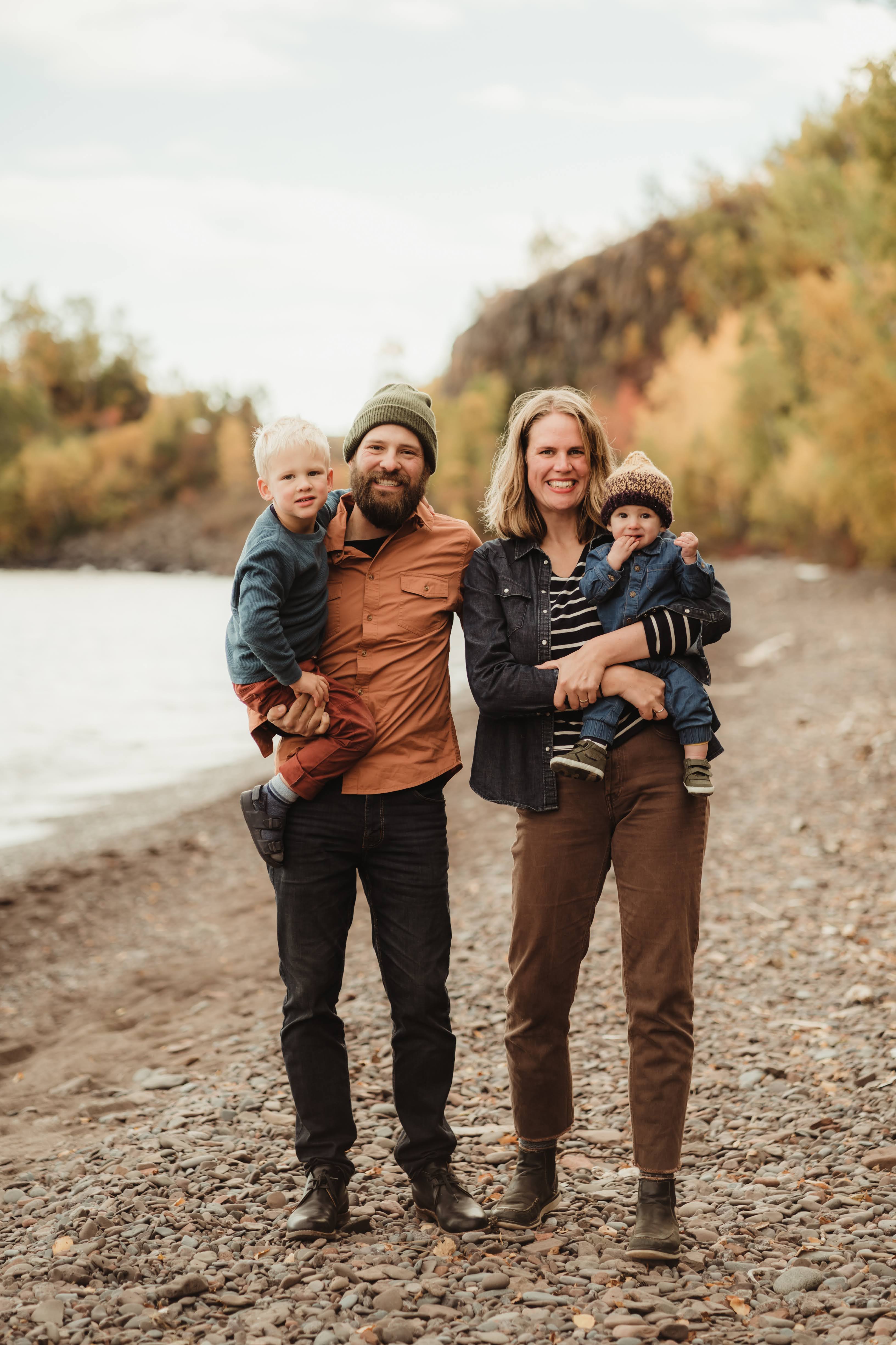 A photograph of Nan Onkka and her family - her husband and two young kids. They are standing along the shores of Lake Superior in the fall. Photo taken by Laura Muus Photography.