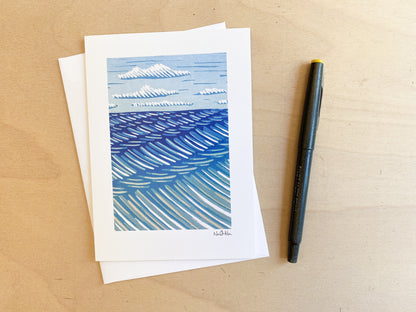 A blank greeting card created by Minnesota artist, Nan Onkka. The original image is a woodcut print depicting a view of Lake Superior. The sky is blue and there are waves on the lake. 